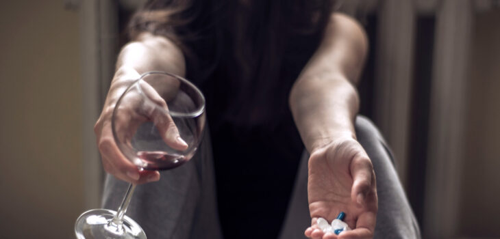 Woman sitting on floor with wine in one hand and benzodiazepines in the other hand.
