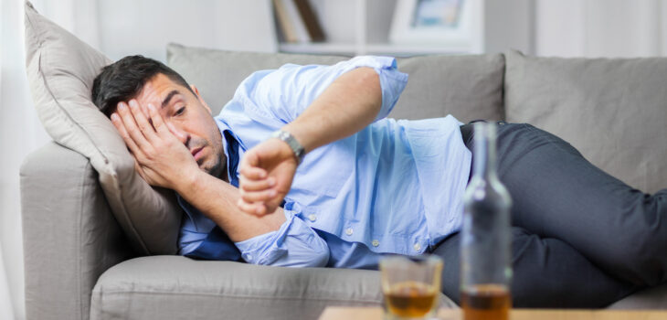 male lying on sofa and looking at wristwatch with alcohol on table
