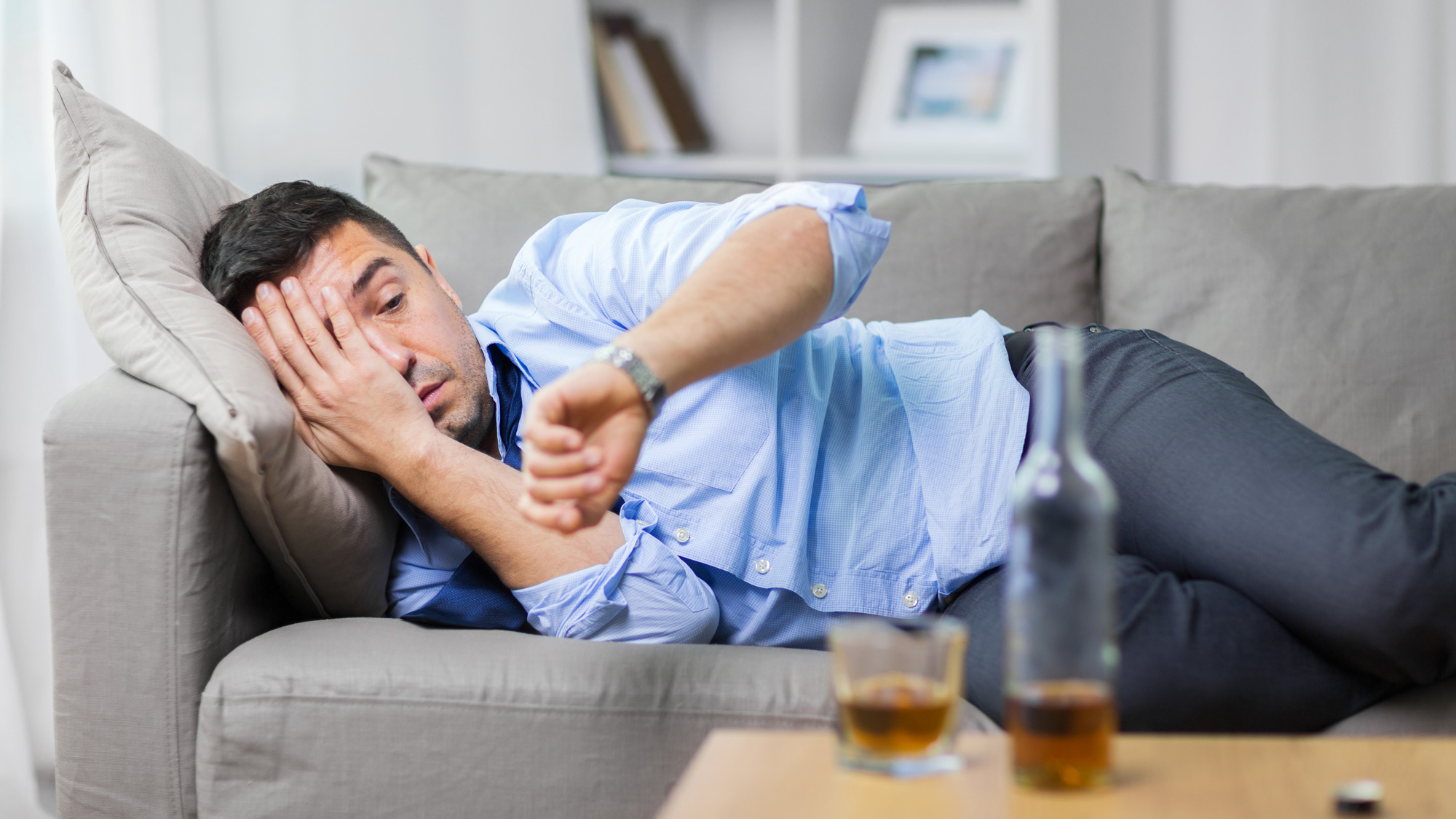 male lying on sofa and looking at wristwatch with alcohol on table